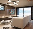 Spacious living room with large windows offering a scenic view, modern lighting fixtures, a beige sofa, a round coffee table, and a wall-mounted TV with various streaming app icons displayed.