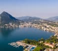 This image shows an aerial view of a scenic lakeside town surrounded by mountains. The town is situated along the shores of a large, blue lake with a marina and boats docked. The town appears to be built on the slopes of the surrounding hills, with a mix