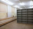 The image shows a large storage room with shelving units along the walls. The room has a concrete floor and fluorescent lighting overhead. There are wooden benches or platforms along one wall, likely used for storage or work. The room appears to be a util