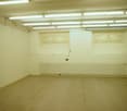 This image shows an empty room with white walls, a tiled floor, and fluorescent lighting fixtures on the ceiling. There are two arched windows on the wall, and a white radiator or heating unit along the wall. The room appears to be a storage or utility sp