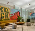 A spacious lobby with yellow and green walls with '5th KLY BECK' on it. A yellow couch, a round table, and round stools with potted plants on it. A glass reception desk with flowers on top.