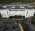 Office building with solar panels, multiple floors, a parking lot, multiple parked cars, and surrounding trees and plants.