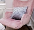 Pink velvet chair with a grey diamond pattern cushion and white wall with radiator. Wooden floor.