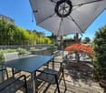 Wooden deck with a table and chairs, umbrella, surrounded by a fence with plants, building and trees in the background