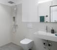 white tile walls, white toilet, white shower, white sink, silver shower head, towel rack, green towel, hand soap, small potted tree