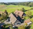 Aerial view of a farm with a main house, outbuildings, cars, and a driveway surrounded by fields and greenery
