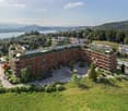 Aerial view of an apartment building with solar panels on the rooftop, situated on a slope with a scenic view of the lake and mountains in the background.