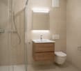 Bathroom with glass walk-in shower, a white toilet, and a wooden cabinet with a sink and faucet, tiled walls, and a wall-mounted mirror with a light.