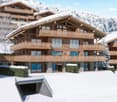 A multi-story chalet with wooden balconies, large windows, surrounded by snow, and a clear view of the mountain range in the background.