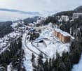 Aerial view of a ski resort with several buildings, including a large building under construction, surrounded by snow-covered trees and mountains.