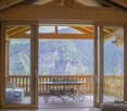 Wooden cabin interior with large windows overlooking a forested mountain landscape, a round wooden dining table with wrought iron chairs on a balcony or terrace