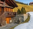 Traditional wooden chalet, wooden balcony, stone wall, snow-covered hill, trees