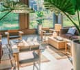 Spacious living room with large windows overlooking a lush, green outdoor area. The room features wooden furniture, including a coffee table, armchairs, and a sofa. Potted plants and decorative items add to the natural, earthy aesthetic.