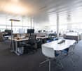 Large open office space with multiple workstations, computers, and a conference table. The space has high ceilings, large windows, and modern office furniture and equipment.