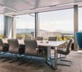 Large conference room with floor-to-ceiling windows offering a panoramic view of the city skyline. The room features a long wooden table surrounded by comfortable leather chairs, and a single blue armchair in the corner. The lighting is provided by recess