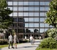 A modern office building with a glass facade surrounded by trees and greenery. People are walking on the path in front of the building.