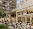 Outdoor dining area with tables and chairs, surrounded by apartment buildings, with a restaurant entrance visible. The area is decorated with plants and flowers.