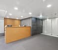 A rectangular reception area with a wooden counter, a black office chair, and gray flooring. To the left, there's a white office chair and a desk with an object. On the right, there's a gray wall with glass doors.