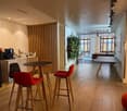 The image shows a modern, open-plan office or coworking space with a coffee and snack bar area. The space features wooden accents, including a wooden divider wall and bar stools, as well as a vertical garden wall. There are various seating options, includ