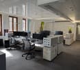 Open office space with white cubicles, computer stations, glass and wooden windows, radiators, central pathway, speakers, and indoor plants.