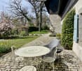 Two-story stone house with a cobblestone path leading to a patio area with wrought iron furniture, surrounded by lush greenery, flowering trees, and a well-maintained garden.