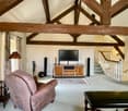 Spacious living room with exposed wooden beams, a large TV console, a plush sofa, and a decorative rug on the floor. The room has a cozy and rustic feel with the wooden elements and the fireplace visible.
