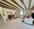 Spacious living room with exposed wooden beams on the ceiling, white walls, hardwood floors, a large L-shaped sofa, an armchair, a coffee table, and a fireplace. The room also has built-in shelves and a staircase leading to another level.