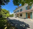2-story house with beige exterior, green shutters, large windows, and a paved driveway or parking area. The house is surrounded by lush greenery and trees.
