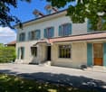 Two-story house with a beige exterior, green shutters, and a tiled roof. The house has several windows and a balcony or terrace on the upper level. The property is surrounded by trees and has a paved driveway or walkway leading to the entrance.