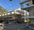 Building under construction with concrete walls, surrounded by metal scaffolding and steel beams, temporary fencing and machinery present, mountain and blue sky in the background.