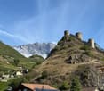 A castle stands on top of a hill overlooking terraced vineyards and a valley with mountains in the background.