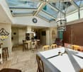 Dining room with wooden tiles, glass roof, chandelier, clock, table, chairs, bar stools, counter, kitchen, fireplace, wall decorations, shelves, microwave