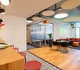 Contemporary styled office room with wooden desk, stools, round dining table, round orange sofa, carpet, plants, LED lights.