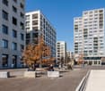 Urban outdoor space between three apartment buildings, surrounded by concrete pathways, benches, trees, and parked bicycles. Several people are seated on benches while others are walking.