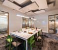 A white conference table, 9 green chairs, carpet, 2 pendant lights, large glass windows.