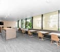 Large open-plan office space with floor-to-ceiling windows providing a view of the surrounding buildings and greenery. The space features a central island-style workstation, comfortable seating areas, and a mix of modern furniture in neutral tones. The fl