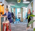 Modern office space with colorful decor, including orange accents, potted plants, and pendant lighting. The space features a kitchen area with a dishwasher, and a man in a suit is working at a desk while other people move through the hallway.