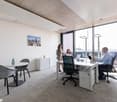 A modern and well-lit office space with white walls, gray carpet, and large windows. The room contains a desk with a laptop, two swivel chairs, and a small round table with two gray chairs. There is a white cabinet and a picture frame on the wall. Two wom