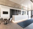 Reception area with reception desk, large monitor, chairs, desk, two people working, bright space, large windows