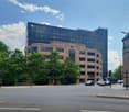 A modern, large office building made of brick and glass stands prominently on a wide street with ample trees, featuring many floors and windows.