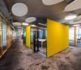 Open office layout with concrete ceiling, yellow accent walls, glass partitions, and carpeted flooring. The space includes various workstations and meeting areas.