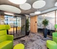 Modern office space with glass-enclosed meeting room, green upholstered chairs and ottomans, large potted plant, and circular lighting fixtures on the ceiling.