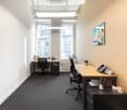 A room with modern minimalist design. There are two desks and chairs facing each other, with a view to the outside of the building. In the center of the room there is a phone and some office materials. The walls are white, and the floor is carpeted.