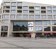 Modern office building with glass windows, curved design, two advertisement banners, main entrance with double doors, red umbrellas at cafe outside