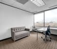 The image shows a small office space with a gray sofa, a desk with a chair, and large windows providing natural light. The room has a neutral color scheme with white walls, gray carpeting, and a suspended ceiling with recessed lighting.