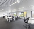 Large open office space with multiple workstations, desks, and chairs. The room has large windows providing natural light and a view of the outdoors. The ceiling is lined with fluorescent lighting fixtures. The floor is covered in dark gray carpeting.