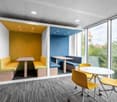 This image shows a modern and colorful office or workspace area. The space features large windows providing a view of the surrounding greenery outside. The room has a mix of seating options, including yellow chairs and a built-in bench with blue cushions.