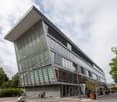 A modern, multi-story building with a unique angular design featuring glass and metal panels. The building has a sleek, contemporary appearance with large windows and a prominent roofline. The surrounding area includes trees and other landscaping elements