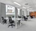 Large open office space with multiple workstations, white tables, black office chairs, and large windows providing natural light. The space has a modern and minimalist design with a mix of gray and orange accents.