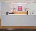 The image shows a modern and minimalist office reception area. The main feature is a large white counter with a wooden base, which appears to serve as the reception desk. On top of the counter, there are various decor items such as plants, a framed artwor